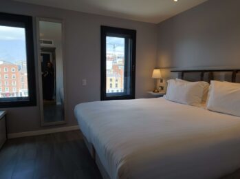 Spacious hotel room with cosy king bed, city view through large windows, elegant decor, bedside lamp, and full-length mirror.