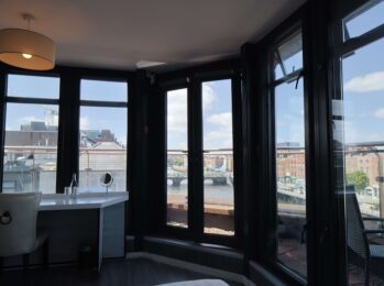 Spacious hotel room with panoramic city views, modern desk, and cosy chair, ideal for relaxation and productivity.
