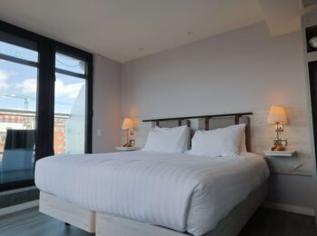 Cosy modern hotel room with a king-size bed, soft lighting, city view from large windows, and ample wardrobe space.