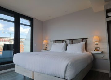 Cosy modern hotel room with a king-size bed, soft lighting, city view from large windows, and ample wardrobe space.