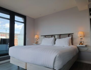 Cosy modern hotel room with a king-size bed, soft lighting, city view from large windows, and ample wardrobe space.
