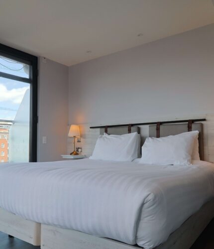 Cosy modern hotel room with a king-size bed, soft lighting, city view from large windows, and ample wardrobe space.