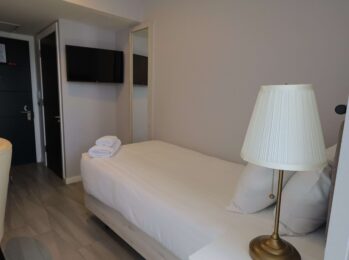 Cozy single hotel room with modern decor, plush bedding, flat-screen TV, lamp, and white towels, ideal for a relaxing stay.